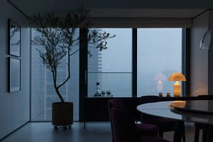 A modern dining room with a round table, purple chairs, and a contemporary lamp. Large windows offer a cityscape view.