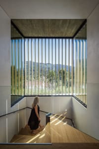 A person walking up a modern staircase with a wooden railing, framed by large vertical windows with views of distant mountain
