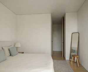 A minimalist bedroom with a bed, lamp, mirror, stool, and rug.