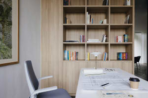 A modern home office with wooden shelves containing books and a white desk with blueprints and a coffee cup on it.