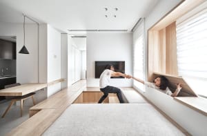 Two people interacting in a modern interior space with wooden benches and white walls, light wood flooring, and a window view