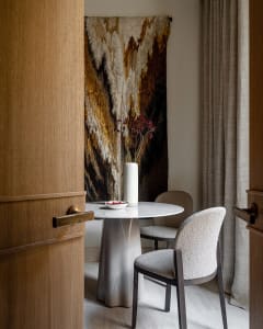 A modern dining room featuring a round marble table with white chairs, a vase with dried flowers, and a textured abstract art