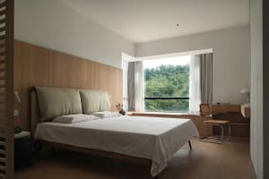 A minimalist bedroom featuring a bed with a wooden headboard and white bedding, surrounded by modern furniture and a large wi