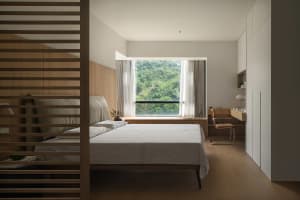A minimalist bedroom featuring a bed with white sheets and a wooden headboard and floor, with a large window showcasing a lus