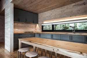 A minimalist kitchen featuring wooden cabinets and countertops, with a long wooden table in the foreground and large windows 