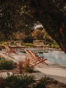 Modern poolside deck featuring wooden lounge chairs on a concrete ground, surrounded by lush greenery and shaded by trees.