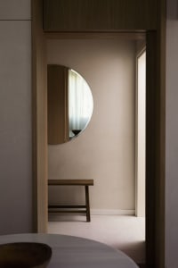 A minimalist bedroom featuring a circular mirror on the wall, a wooden bench, and sheer curtains, creating a serene and moder