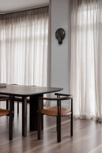 A modern dining room featuring wooden furniture, leather seating, light-colored sheer curtains, and wall-mounted light fixtur