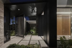 A modern entrance with a black metal grid door, illuminated at night with surrounding greenery and concrete paving.