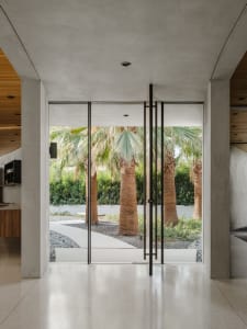 Modern entryway with glass doors, concrete interior, and a garden featuring palm trees.