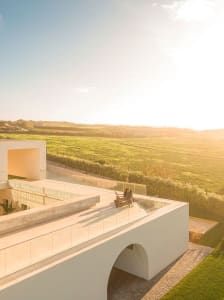A modern residential veranda with concrete flooring and glass railings overlooking green fields at sunset.