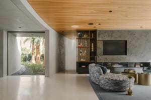 A modern living room with concrete walls, a white floor, a wooden ceiling, and a view of palm trees.