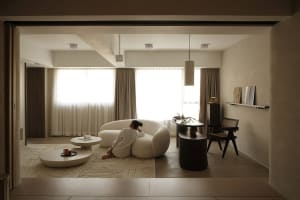 A modern living room featuring a cozy, white, round sofa with a woman sitting on it, accompanied by a coffee table and variou