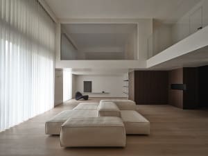 A modern minimalist living room with large windows and a wooden floor, featuring a beige modular sofa and a glass partition.