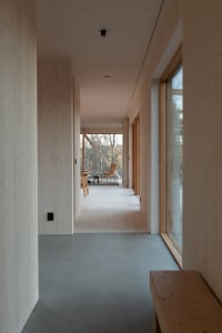 A modern minimalist hallway featuring wooden floors and walls, with a view of an outdoor seating area through large windows.