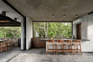 A modern kitchen with exposed concrete ceiling, wooden stools, and a view of trees through large windows.