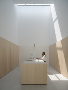 Person preparing food in a minimalist modern kitchen with wood cabinets, white countertops, and overhead lighting.