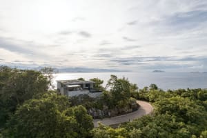 Modern architectural home with concrete walls and a glass terrace overlooking the ocean from a rocky cliffside.