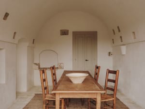 A rustic dining room with a wooden table, chairs, and a central bowl on the table. The walls are made of drywall with visible