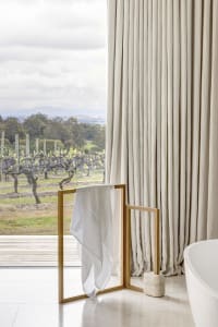 A spacious modern bathroom with long beige curtains, a towel rack, a bathtub, and a view of a vineyard.