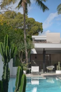 Modern minimalist poolside area with concrete and glass materials, featuring a residential home and outdoor seating area.