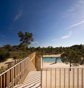 A minimalist outdoor swimming pool with concrete edges and wooden fences, surrounded by natural vegetation under a clear blue