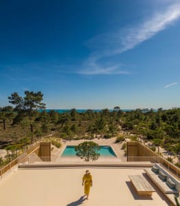 A minimalist patio area with a pool, sand, and wood accents, overlooking a serene coastal landscape.