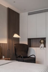 Modern bedroom with a dark wooden accent wall, a dark armchair, a floor lamp, and a white bed with striped sheets.