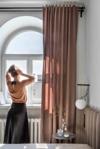 A person standing by a large arched window with textured curtains, looking at the view outside.