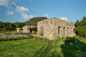 A modern stone house with a peaceful, green landscape and a forested hill in the background.