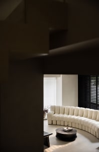 A modern living room featuring a circular sofa with plush cushions, a round coffee table, and black window shutters. The spac