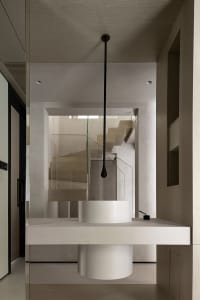 A modern minimalist bathroom with a white floating sink and a glass-enclosed staircase in the background.
