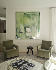 A modern living room with green velvet armchairs, a marble coffee table, and a large abstract green painting.