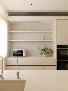 A modern, minimalist residential kitchen with white cabinetry, shelves, and appliances, featuring a vase of flowers and a mic