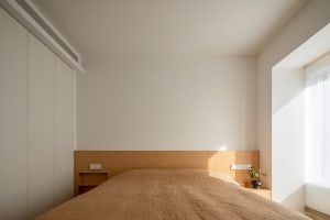 Minimalist bedroom featuring a wooden bed with a plain wall and a window with blinds, highlighting a serene and modern aesthe