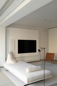 A modern minimalist living room featuring a white chaise lounge, a flat-screen TV mounted on the wall, and a simple, elegant 