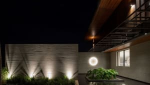 A modern residential architecture featuring concrete walls, illuminated by warm lights, and adorned with green plants in plan
