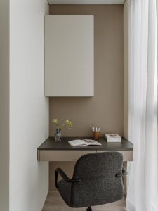 A minimalist home office featuring a beige-grey desk with a wooden drawer, a grey upholstered chair, and a beige-grey cabinet