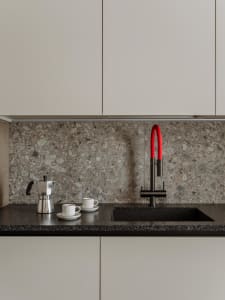 A sleek modern kitchen featuring a black terrazzo countertop with a black and red faucet, white espresso cups, and a silver e