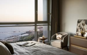 Modern minimalist bedroom with a bed, chair, and table by a large window overlooking the ocean.