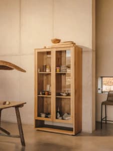 A minimalist storage cabinet with glass doors placed against a concrete wall, containing various dishes and jars.
