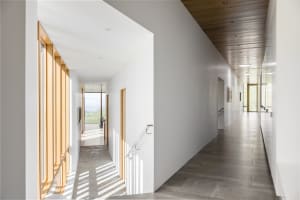 A minimalist hallway with white walls, wooden ceiling, concrete flooring, and large windows providing natural light.