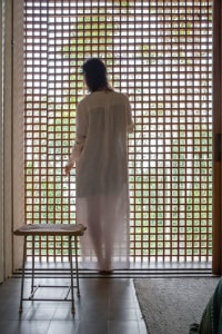 A woman is standing in front of a wooden lattice window with a stool in the foreground and a textured rug on the floor.
