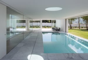 Modern residential swimming pool with clear glass walls and a view of the outdoor area with greenery and a round sofa.