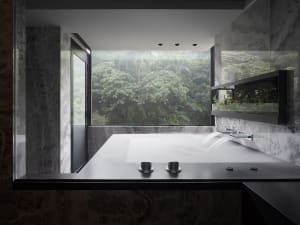 Modern minimalist bathroom with marble fixtures, large window overlooking greenery, and water feature.