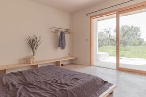 A minimalist modern bedroom with a concrete floor and wall, featuring a bed with a gray blanket and a small plant.