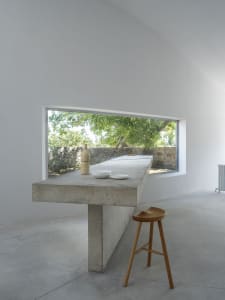 A minimalist concrete table with a window displaying greenery, a stool, and a vase.