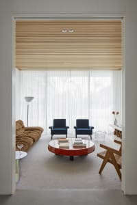 A modern living room with transparent windows, featuring two blue armchairs, a brown sofa, a round coffee table, and a white 