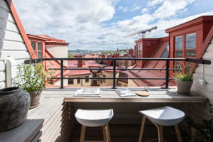 A balcony with wooden railings offering a view of rooftops and the cityscape below, with potted plants and seating.
