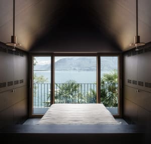 Minimalist bedroom featuring a bed with a white sheet against a window with a scenic view of mountains and a lake.
